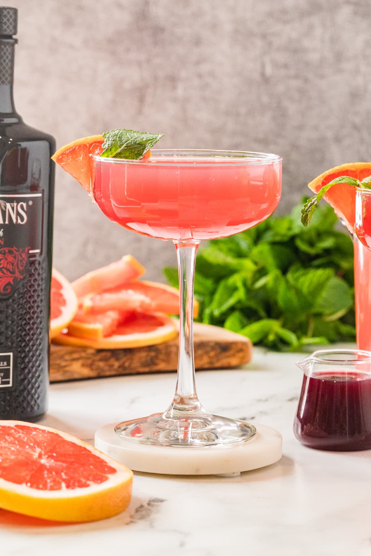 Finished grapefruit and pomegranate cocktail garnished with mint and grapefruit. A pink grapefruit and pomegranate cocktail served in a coupe glass, garnished with a grapefruit slice and mint, with fresh citrus and syrup in the background.