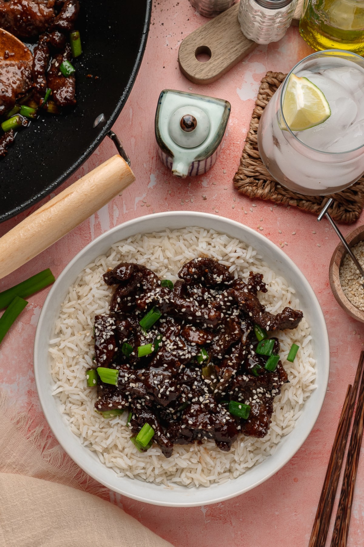 A bowl of saucy sliced beef served over white rice and finished with green onions and sesame seeds on the table.
