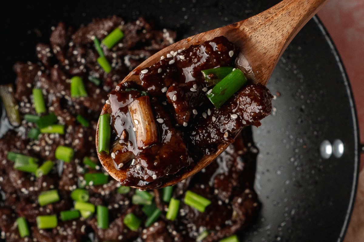 A wooden spoon lifting saucy slices of beef with green onions and sesame seeds from a hot skillet.