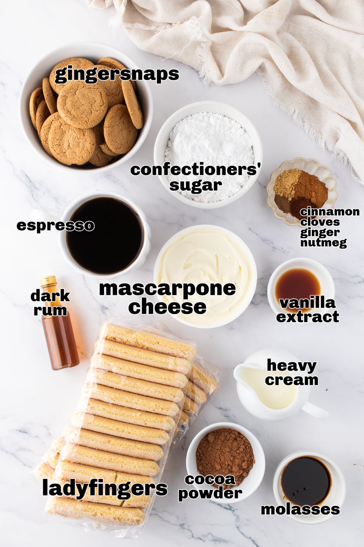 A top-down view of the gingerbread tiramisu ingredients laid out on a marble surface, including gingersnaps, ladyfingers, mascarpone, espresso, spices, molasses, and confectioners’ sugar.