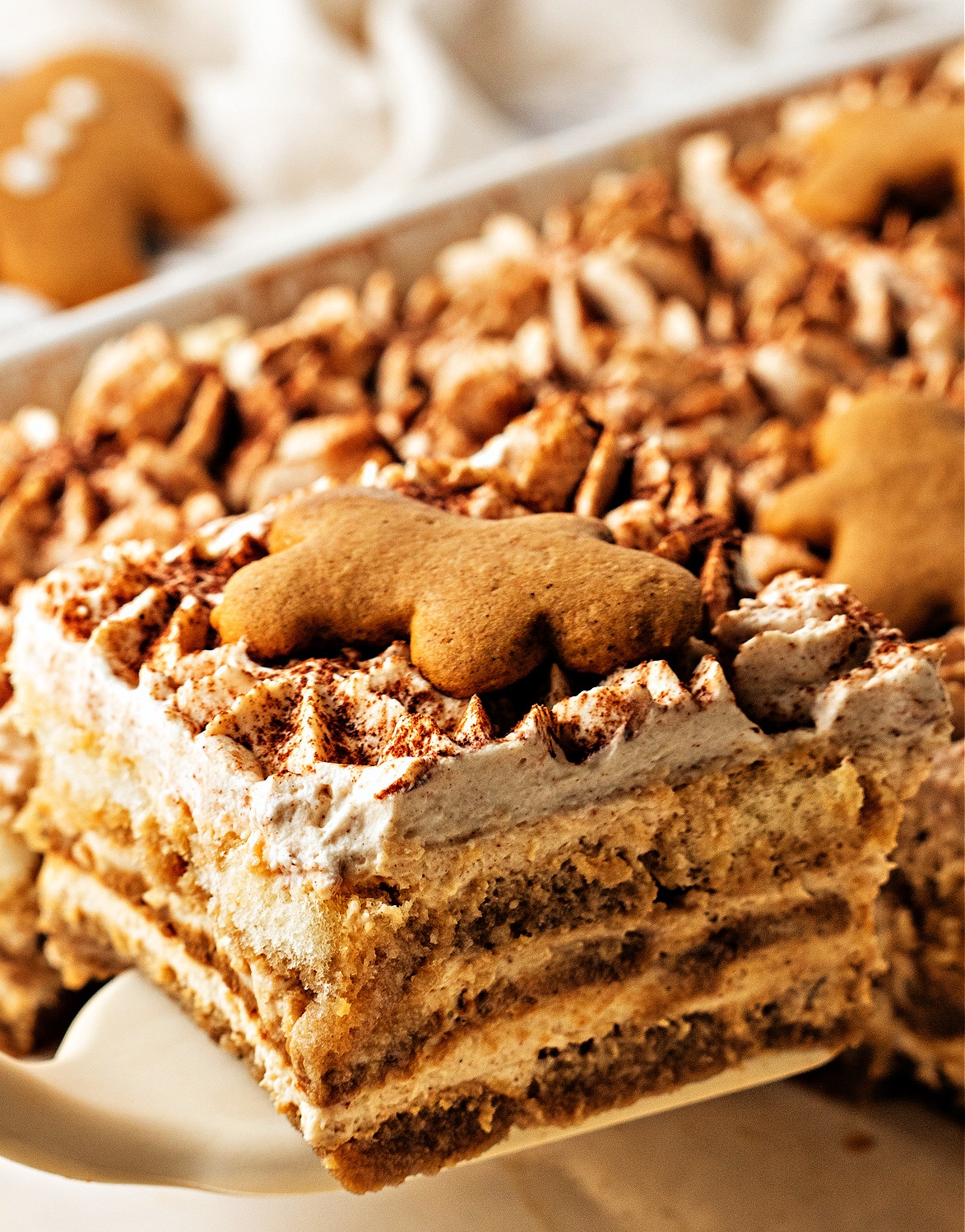 A close slice of gingerbread tiramisu lifted from the pan, showing soft espresso-soaked layers, spiced mascarpone cream, and a small gingerbread cookie resting on top.