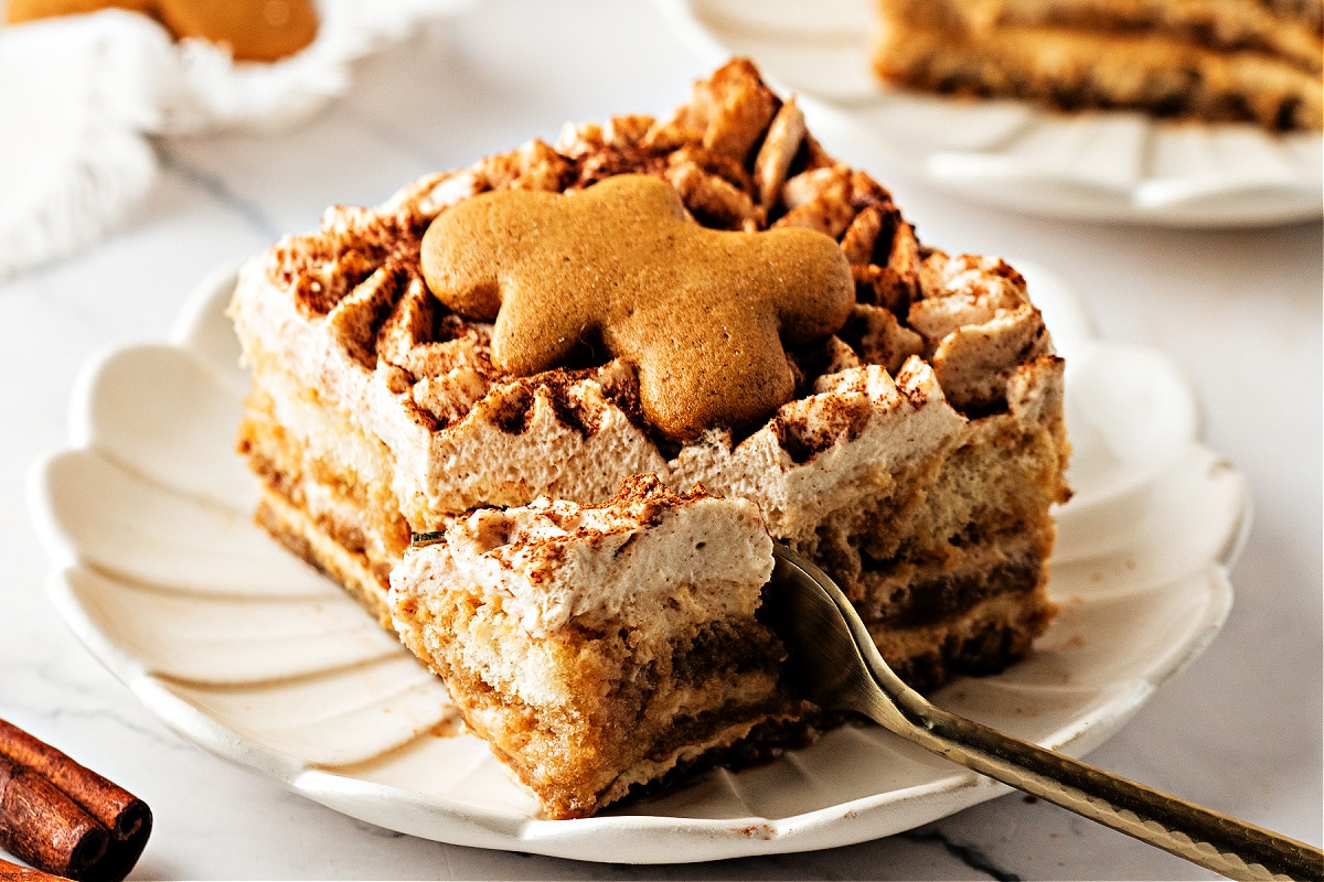 A slice of gingerbread tiramisu with a gingerbread cookie on top, showing the creamy spiced layers and a fork taking the first bite on a scalloped plate.