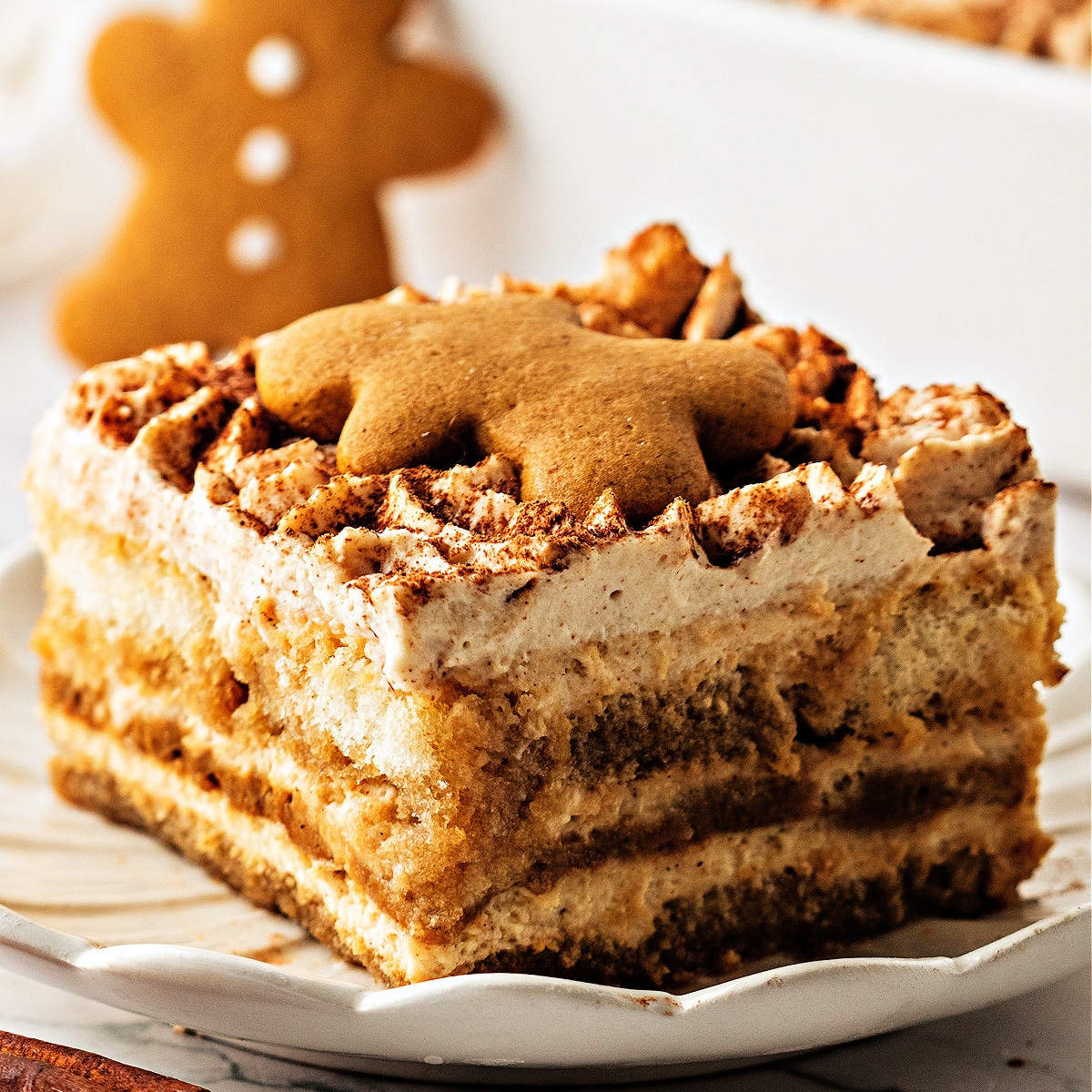 A close-up slice of layered gingerbread tiramisu topped with a small gingerbread cookie, showing the soft espresso-soaked ladyfingers and spiced mascarpone cream.