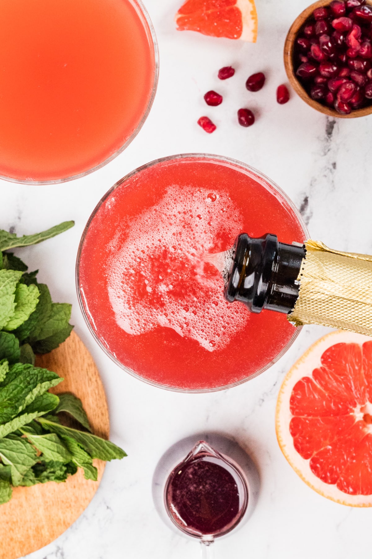 Top-down view of a grapefruit pomegranate gin fizz with prosecco being poured. Top-down view of a grapefruit pomegranate gin fizz being topped with prosecco, showing fine bubbles forming over the pink cocktail with fresh grapefruit, pomegranate arils, and mint nearby.