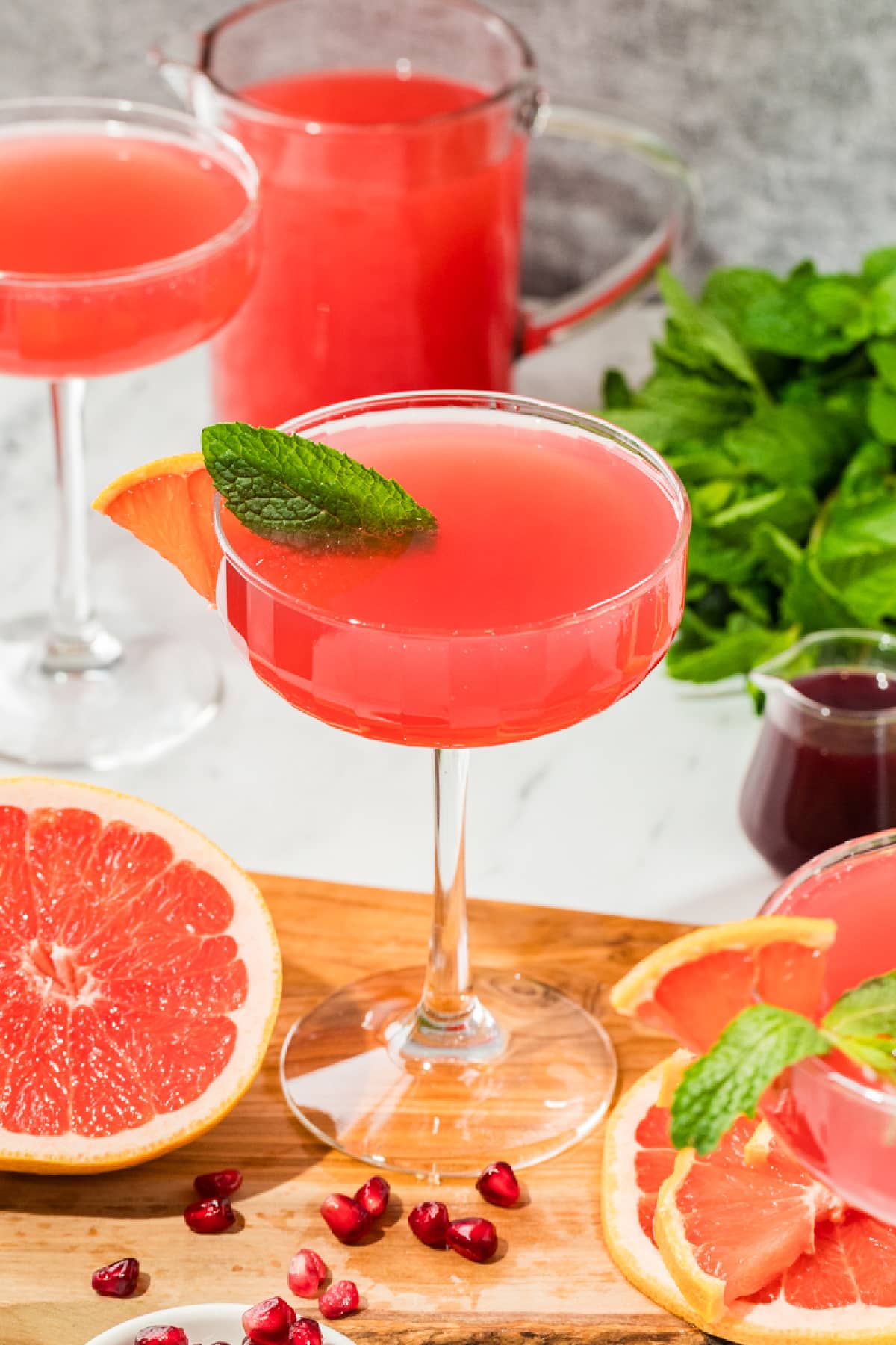 Grapefruit and pomegranate cocktails arranged with fresh citrus and mint for brunch. Coupe glasses filled with a pink grapefruit and pomegranate cocktail on a wooden board, surrounded by fresh grapefruit halves, pomegranate arils, mint, and a pitcher of juice.