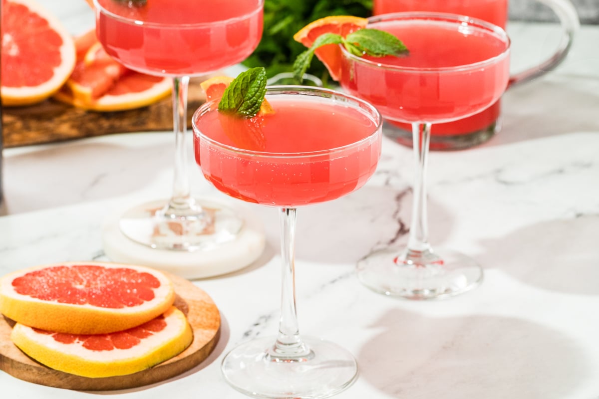 Pink grapefruit and pomegranate gin cocktails served in coupe glasses for brunch. Coupe glasses filled with a pink grapefruit and pomegranate gin cocktail, garnished with fresh grapefruit slices and mint on a marble surface.