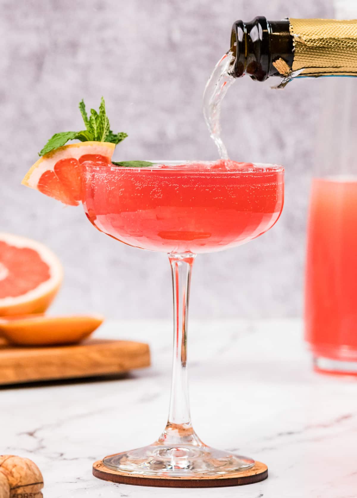 A grapefruit pomegranate gin fizz topped with prosecco in a coupe glass. A grapefruit pomegranate gin fizz being topped with prosecco in a coupe glass, showing the deep pink color, fine bubbles, and a garnish of grapefruit and mint.