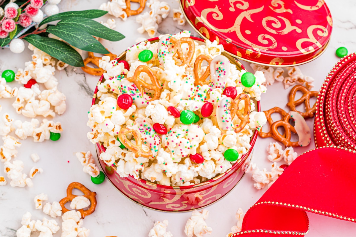 This festive popcorn mix is styled in a holiday tin with sprinkles and candy. Festive popcorn mix with white chocolate–dipped pretzels, red and green candies, and sprinkles in a holiday tin.