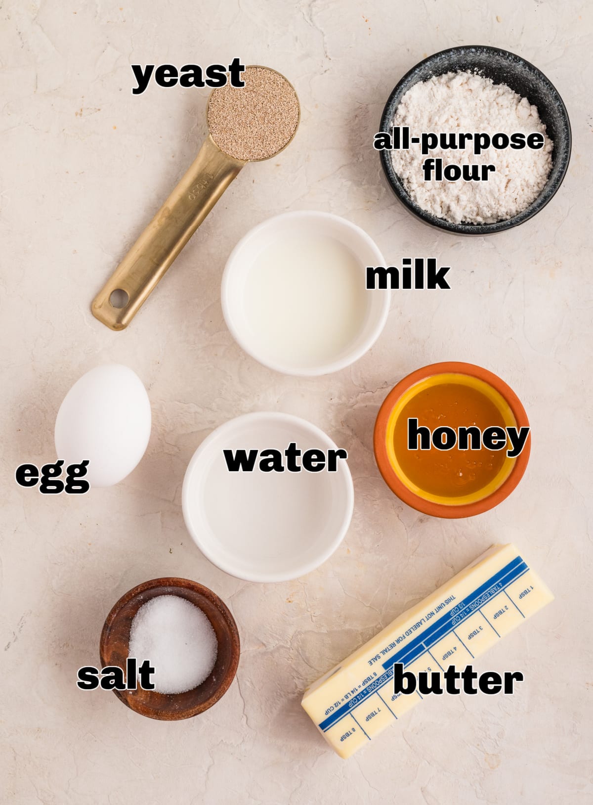 An overhead layout of the dough ingredients for pull-apart rolls, with flour, yeast, milk, water, honey, egg, salt, and butter clearly arranged and labeled. Measured ingredients for pull-apart rolls including flour, yeast, milk, water, honey, egg, salt, and butter.