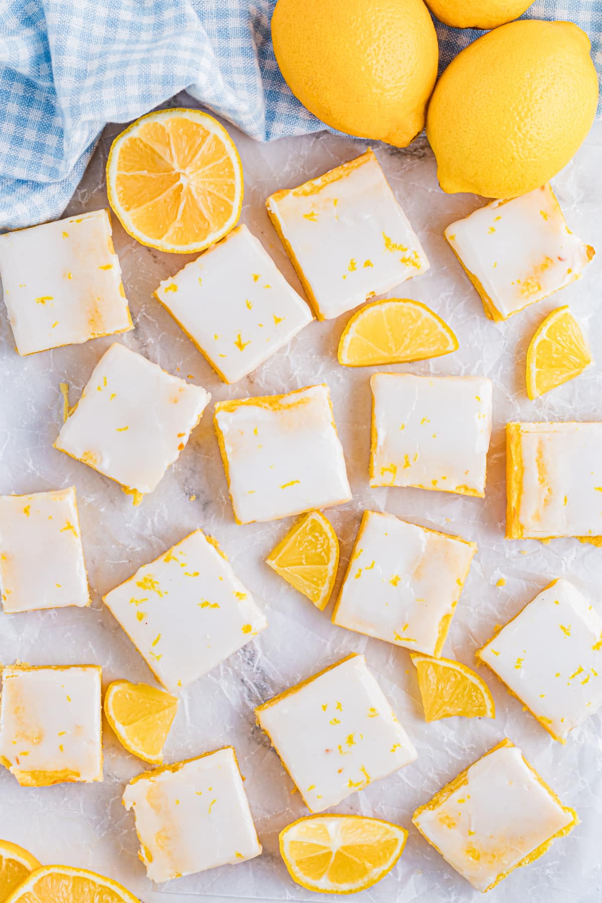 Glazed lemon dessert squares scattered with lemon wedges on parchment paper. Lemon blondies arranged across parchment with fresh lemon slices, showing the glazed tops and clean, square cuts.