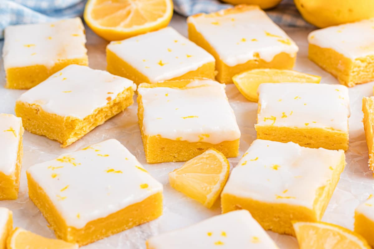 Finished lemon blondies styled with whole lemons and parchment for a clean, bright presentation Lemon blondies arranged in a loose grid on parchment with fresh lemons nearby, highlighting the pale glaze and bright citrus color.