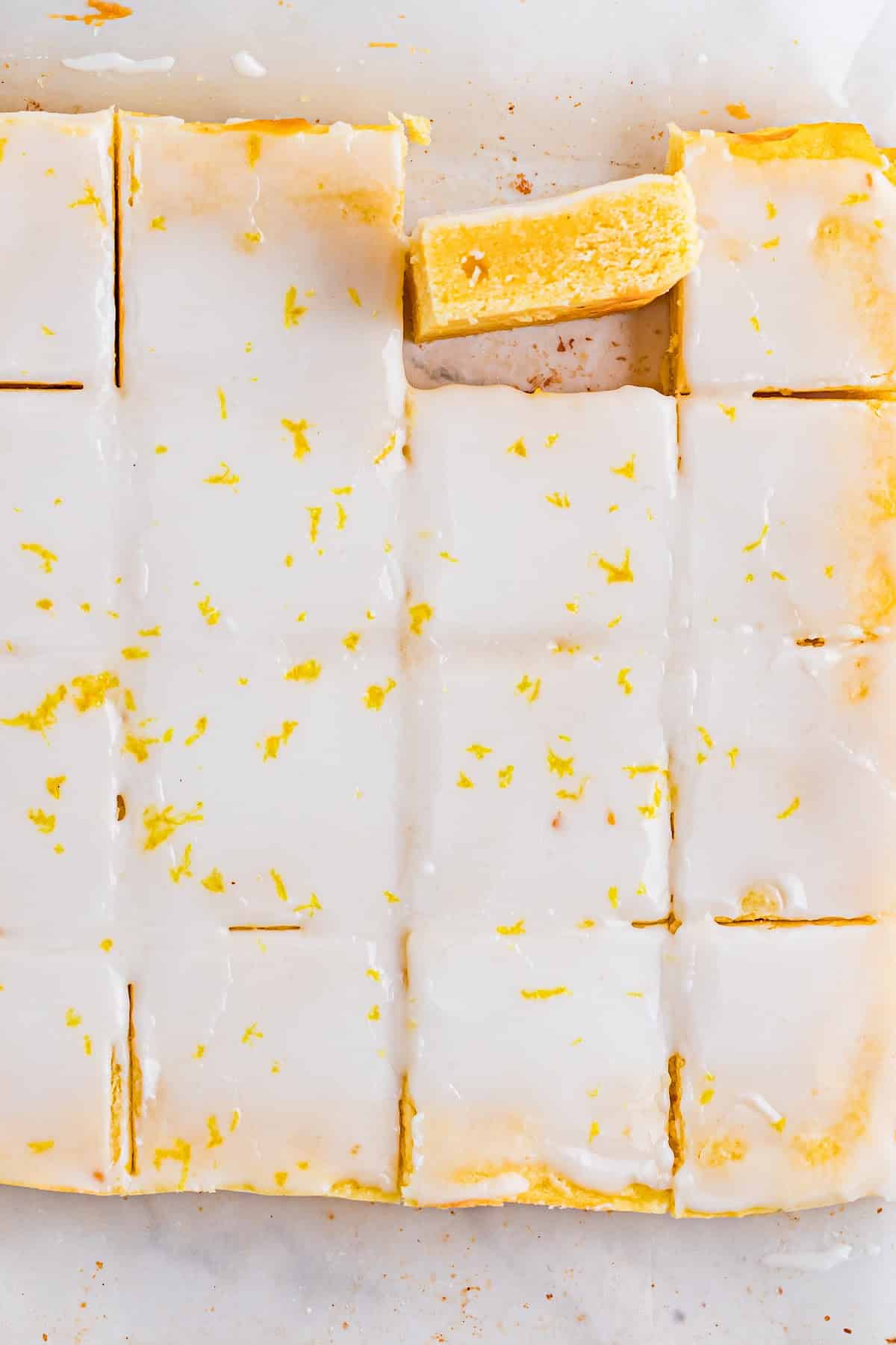 Fully baked blondies sliced in the pan with a set lemon glaze and visible citrus crumb. Overhead view of sliced lemon blondies in the pan, showing a smooth lemon glaze, clean cuts, and a dense yellow crumb.