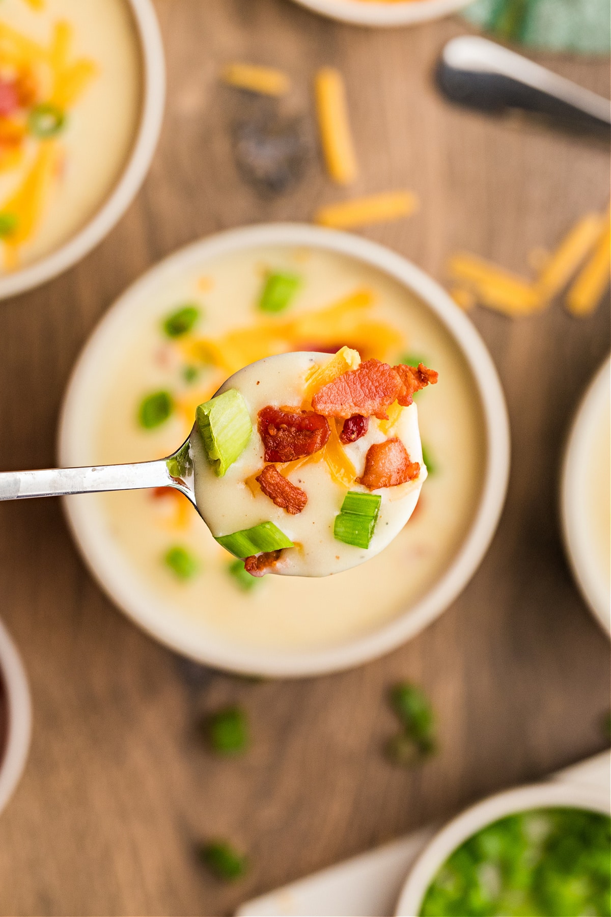 A close-up spoonful of creamy potato soup finished with bacon, cheddar, and scallions. A spoonful of creamy potato soup topped with crisp bacon, melted cheddar, and chopped scallions held over a bowl.
