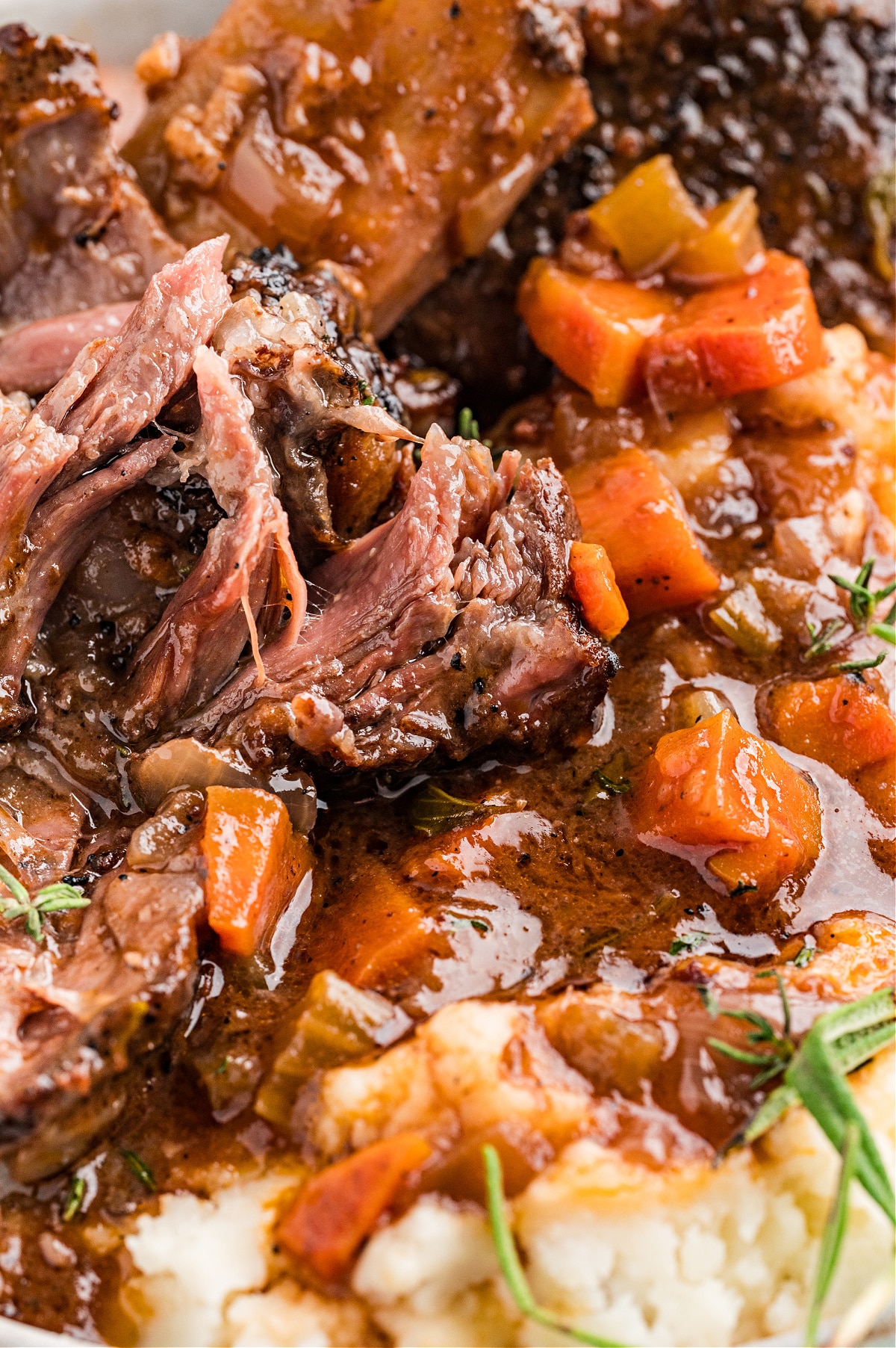 Fork-tender braised short ribs in thick sauce with softened vegetables and herbs. Close-up of oven-braised short ribs pulled apart in a rich sauce with carrots and herbs, showing the tender texture after long cooking.