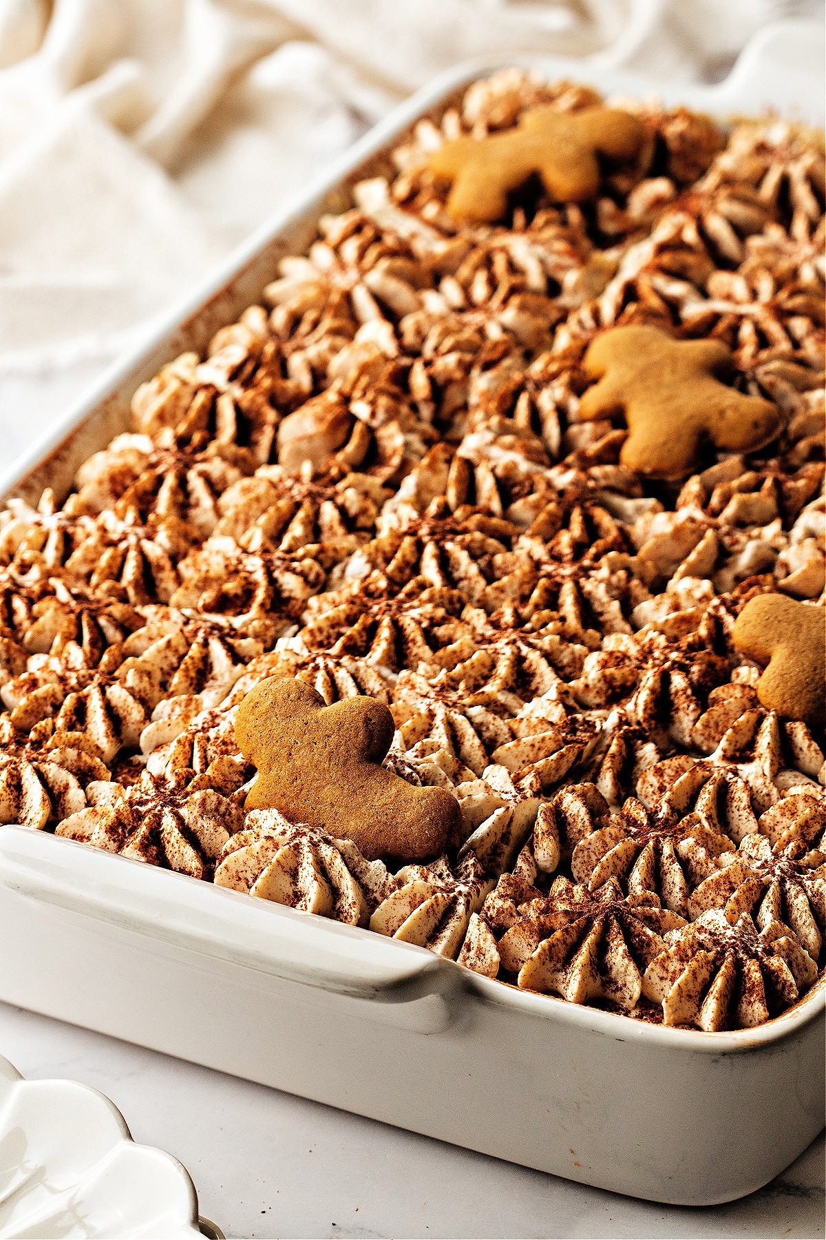 A full pan of spiced holiday tiramisu topped with piped cream, cocoa dusting, and gingerbread men resting across the surface.