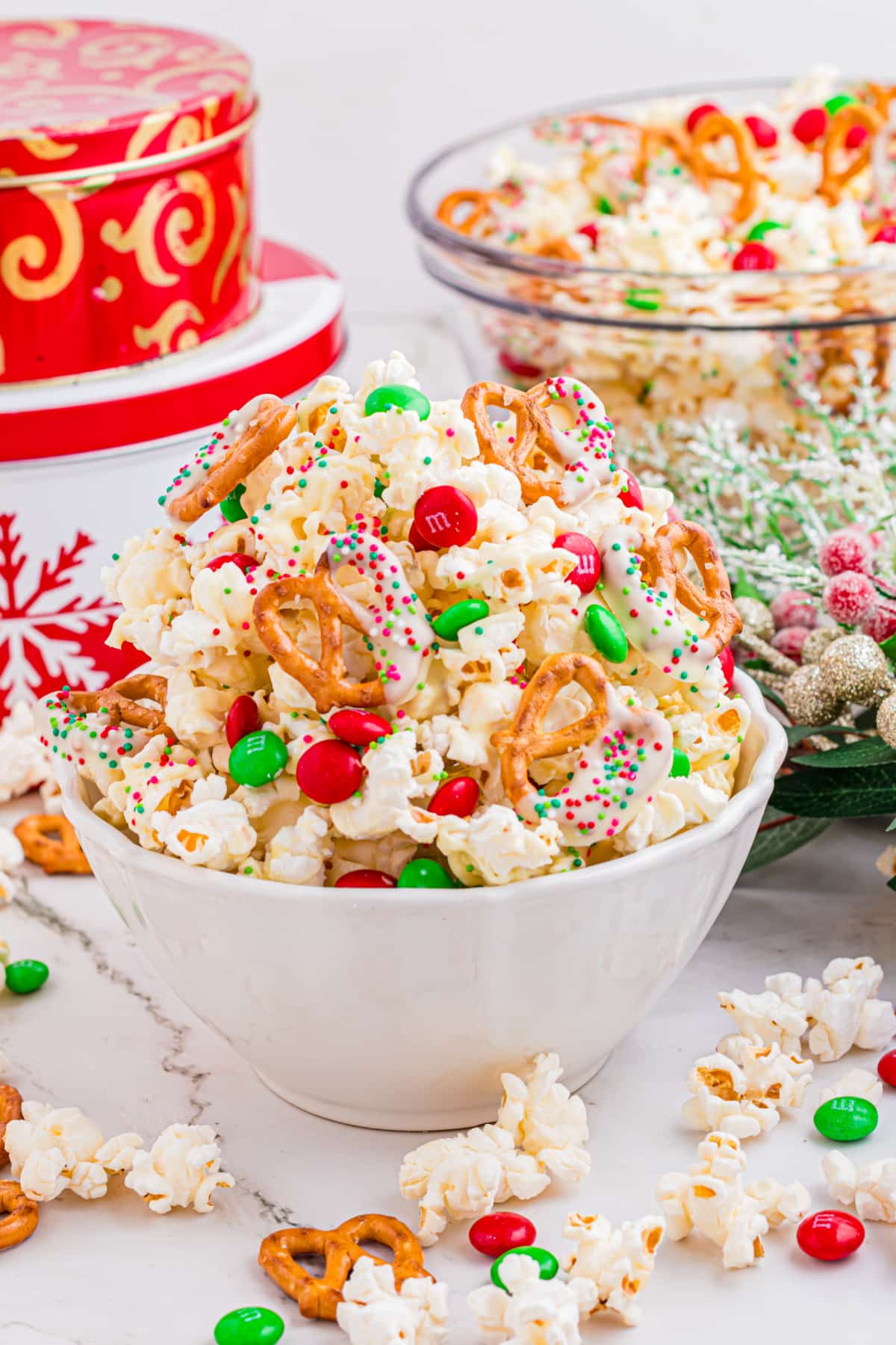 This Christmas popcorn mix is served in a white bowl ready for sharing. Christmas popcorn mix piled into a white bowl with white chocolate–dipped pretzels, red and green candies, and holiday sprinkles.