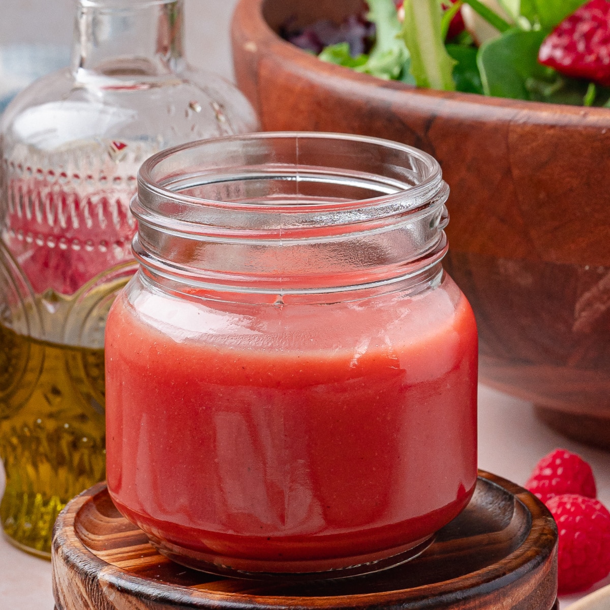 A small jar of homemade raspberry dressing resting on a wooden coaster with a salad bowl blurred in the background.