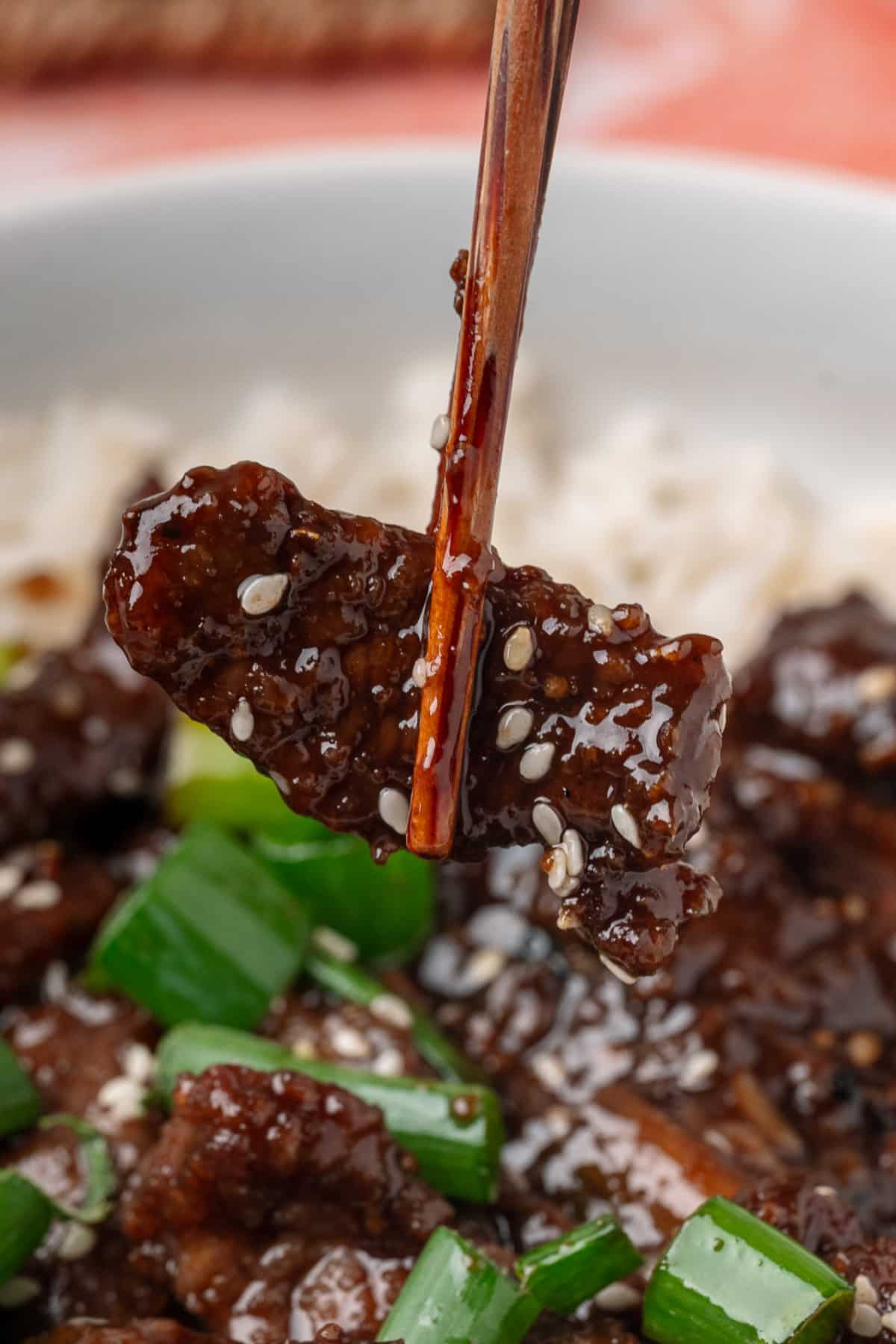 Ginger beef held with chopsticks, coated in a glossy soy ginger sauce with sesame seeds and green onions.