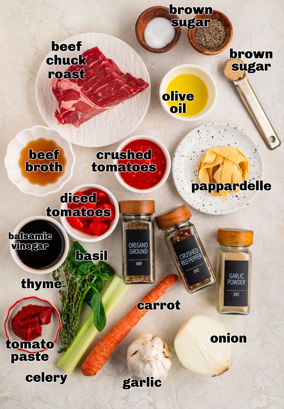 Ingredients for an oven-braised beef ragù laid out on a counter, including chuck roast, tomatoes, herbs, aromatics, oil, seasonings, and pappardelle.