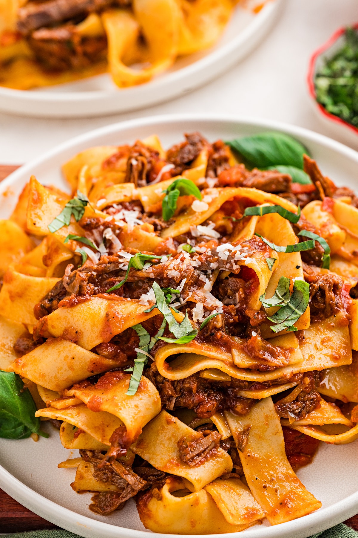 A close-up plate of slow-braised beef ragù with pappardelle, showing wide ribbons of pasta tangled with tender shredded beef, tomato-rich sauce, fresh basil ribbons, and a light grating of parmesan.