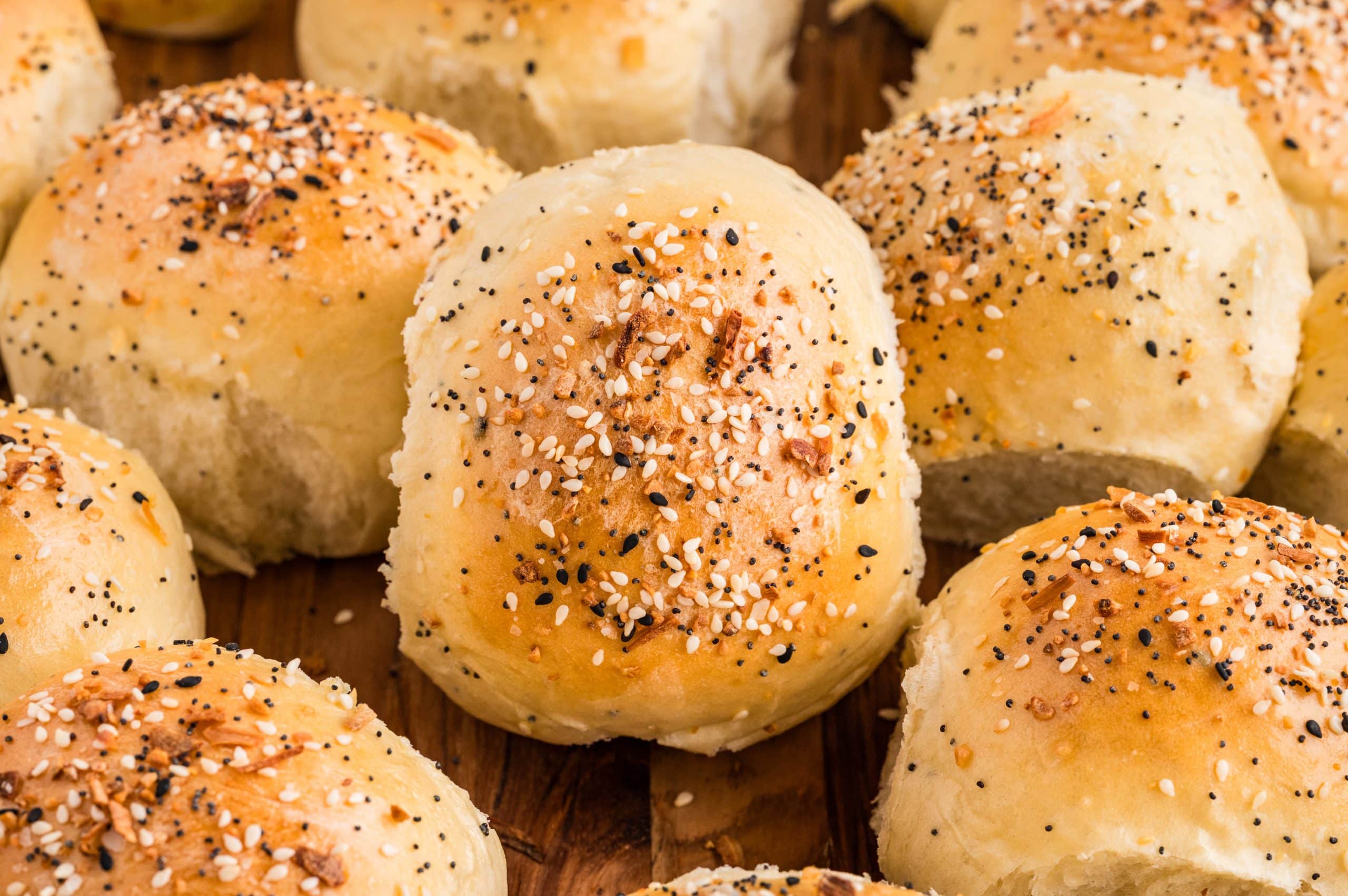 Golden baked rolls arranged on a wooden board, topped with sesame seeds, poppy seeds, garlic, and onion, with one roll split open to show the soft interior.