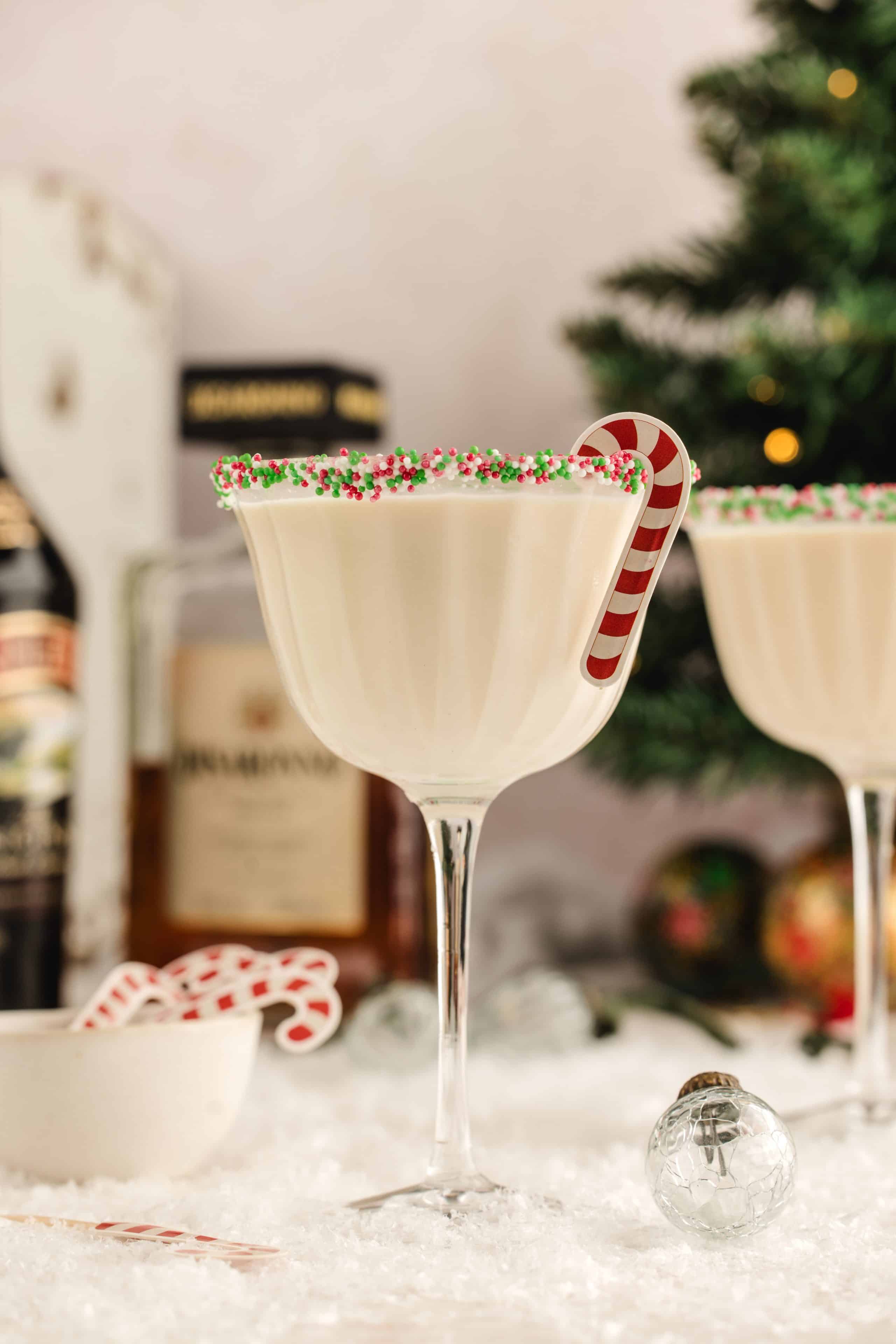 A festive cream cocktail with a candy cane garnish in a sprinkle-rimmed coupe. A creamy holiday cocktail in a sprinkle-rimmed coupe with a candy cane garnish, surrounded by snowy décor and bottles in the background.