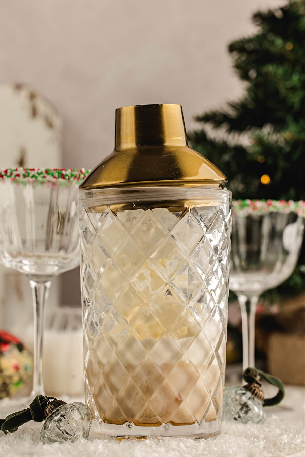 Cream liqueur poured over ice in a mixing glass to make a festive holiday cocktail. Irish cream being poured from a gold jigger into a mixing glass filled with ice and amaretto, with sprinkle-rimmed holiday coupes in the background.