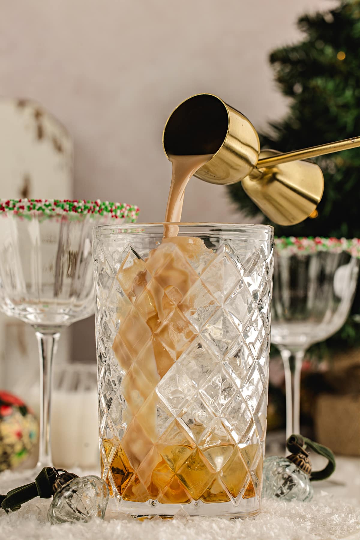 Cream liqueur poured over ice in a mixing glass to make a festive holiday cocktail. Irish cream being poured from a gold jigger into a mixing glass filled with ice and amaretto, with sprinkle-rimmed holiday coupes in the background.