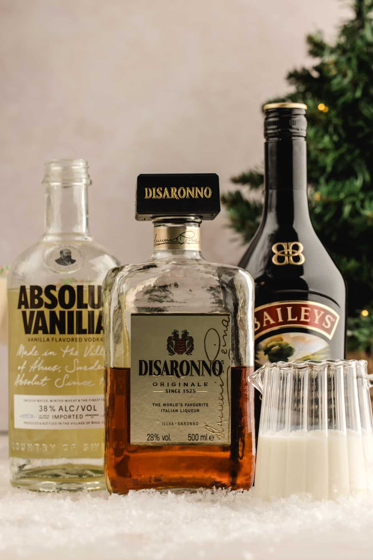 Holiday martini ingredients including vanilla vodka, amaretto, Irish cream, and cream on a snowy tabletop. Vanilla vodka, amaretto, Irish cream, and a small pitcher of cream arranged together on a snowy surface with a Christmas tree blurred behind them.