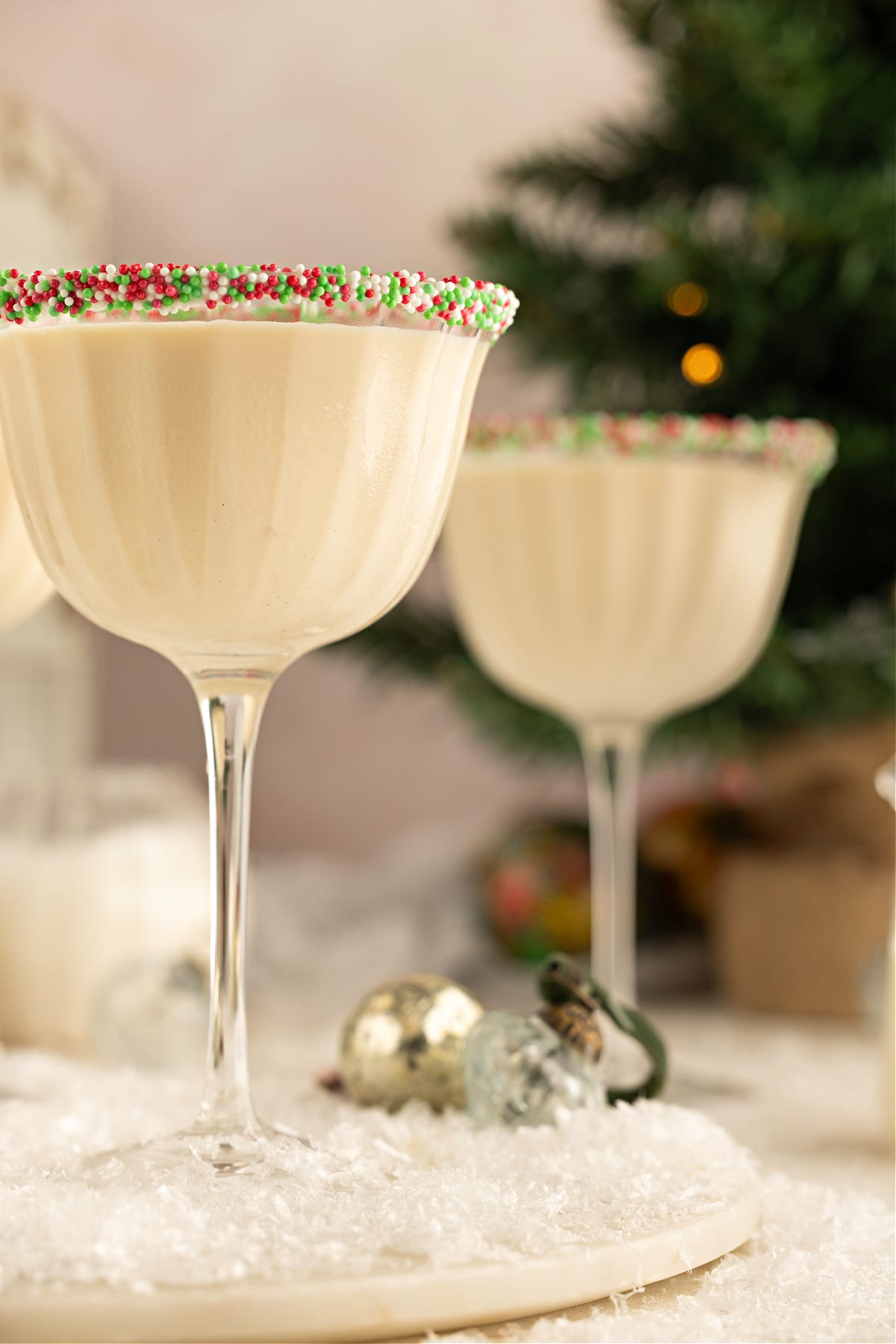 A Sugar Cookie Martini in a sprinkle-rimmed coupe set on a snowy holiday scene. A Sugar Cookie Martini in a scalloped coupe glass with a holiday sprinkle rim, sitting on a snowy tabletop with blurred Christmas ornaments and a tree in the background.