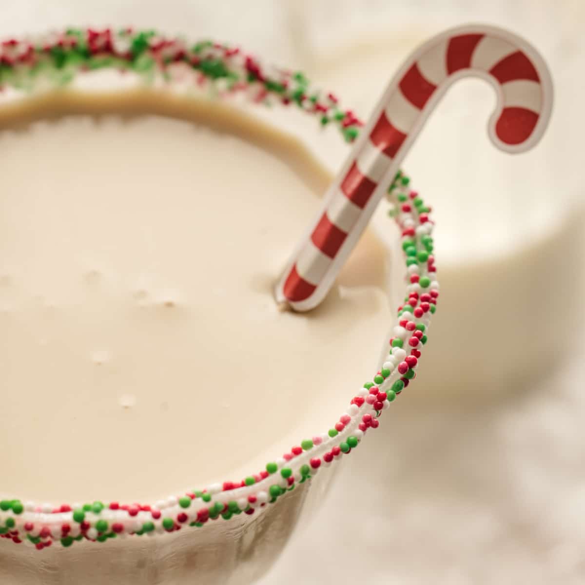 A close-up of a holiday cocktail rimmed with red, green, and white sprinkles, finished with a tiny candy cane stir stick resting on the creamy surface.