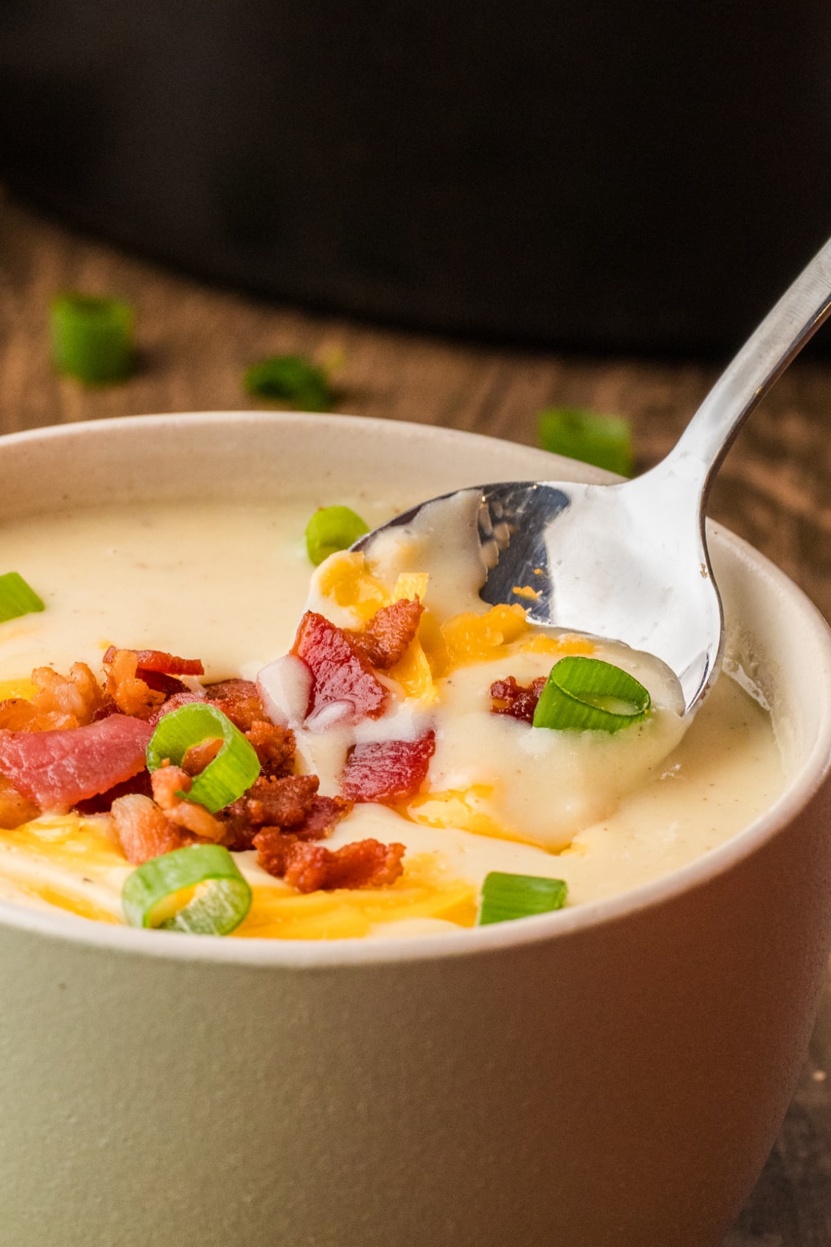 A close-up spoonful of loaded baked potato soup with bacon, cheddar, and scallions. A spoon dipping into loaded baked potato soup, showing chunks of potato, melted cheddar, crisp bacon, and scallions in a creamy base.