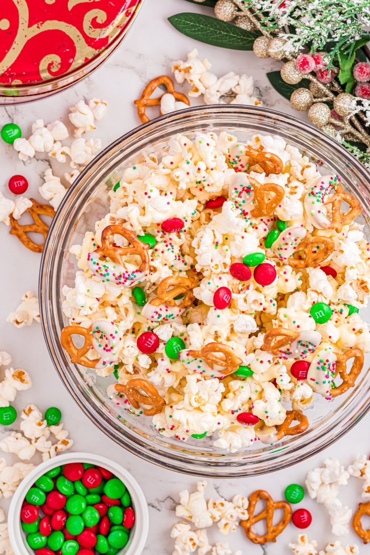 A bowl of white chocolate popcorn mixed with pretzels and holiday candy sits on the counter. White chocolate popcorn with dipped pretzels, red and green candy-coated chocolates, and holiday sprinkles in a glass bowl.
