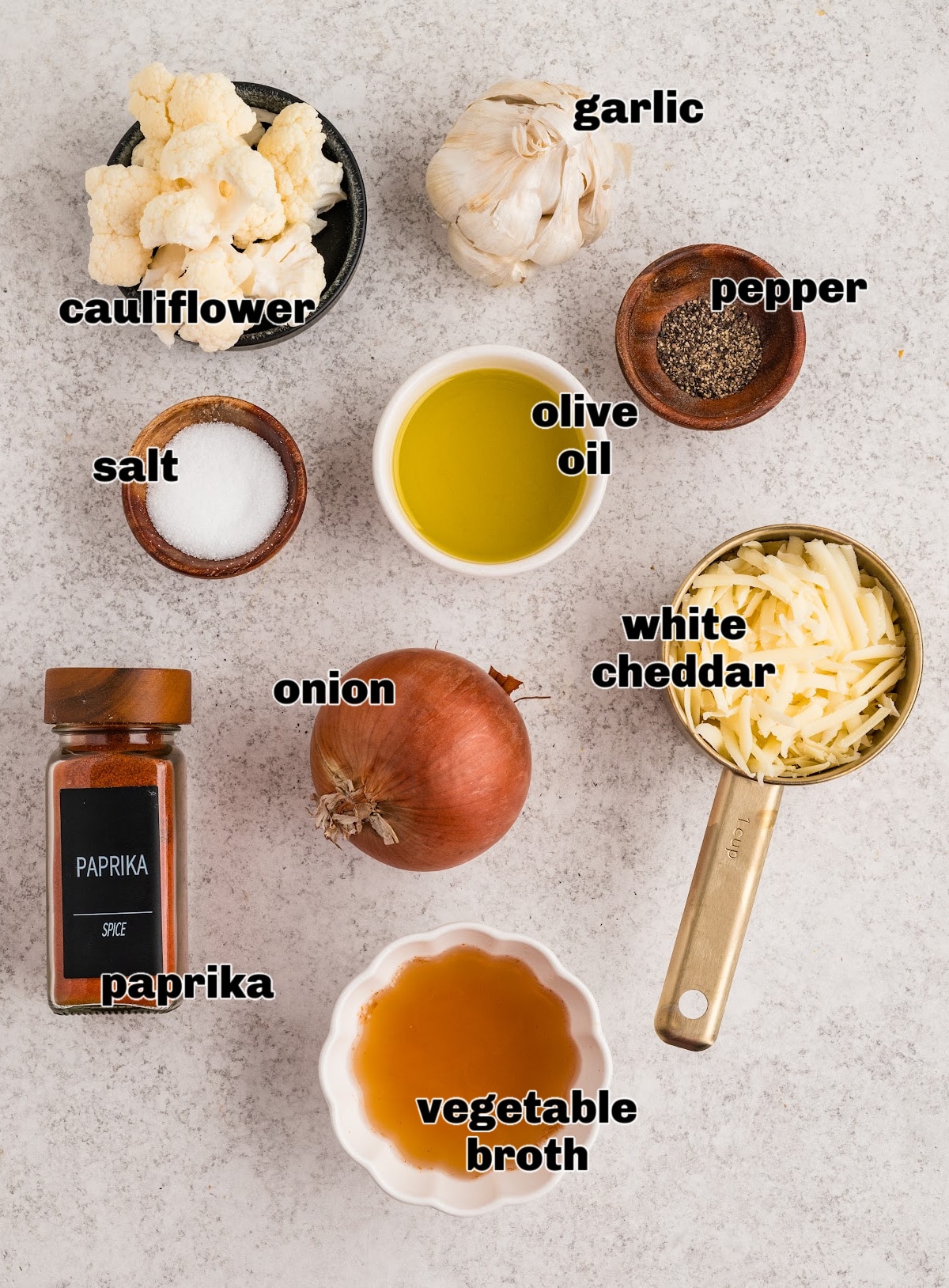The ingredients used to make this cauliflower soup laid out before cooking, including cauliflower, garlic, onion, olive oil, broth, and white cheddar. Ingredients for cauliflower soup arranged on a light surface, including cauliflower florets, garlic, onion, olive oil, vegetable broth, white cheddar, salt, pepper, and paprika.