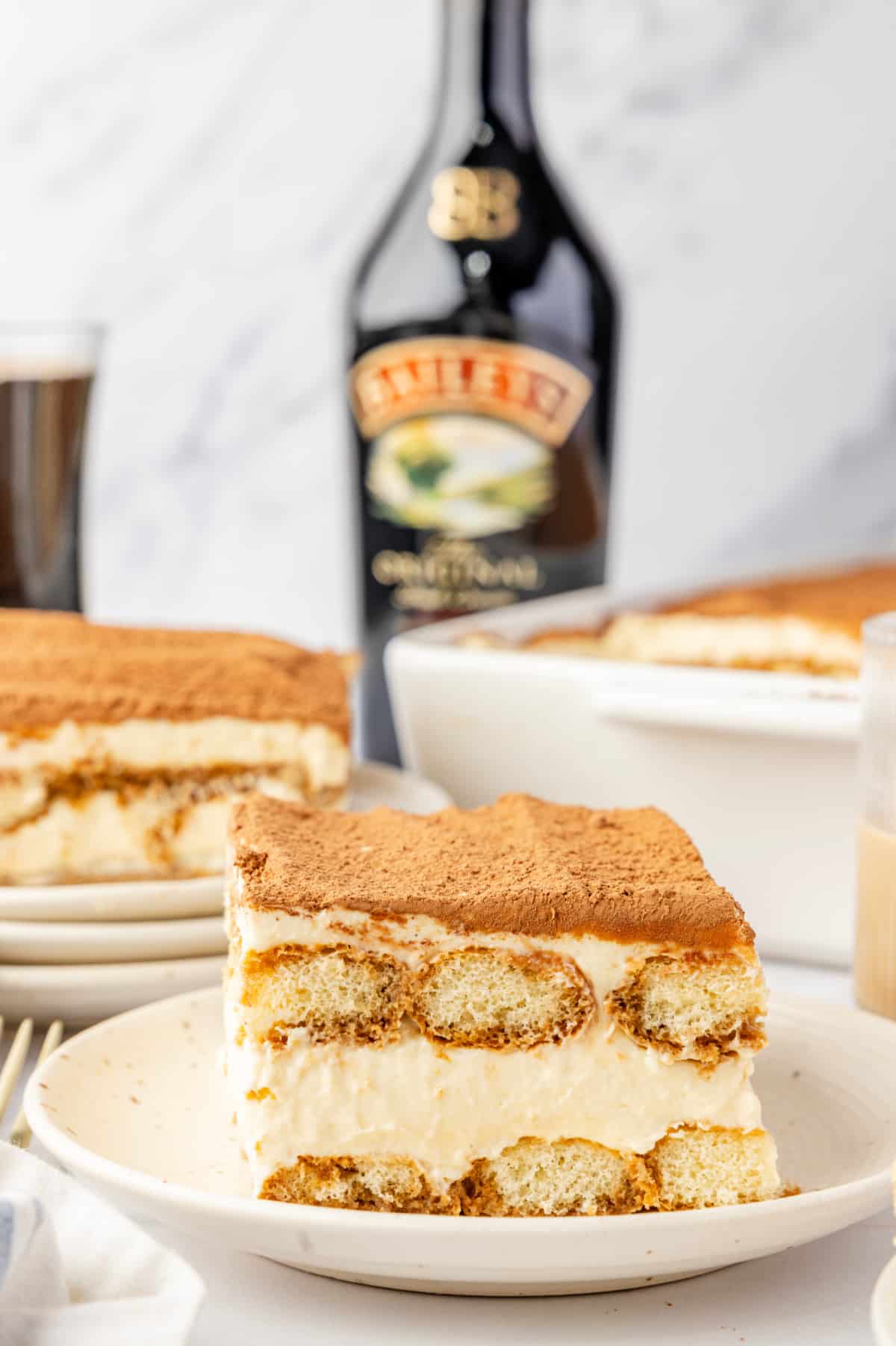 A plated tiramisu slice with cocoa on top, layered ladyfingers and mascarpone cream, and Baileys Irish cream set behind it. Plated slice of tiramisu with visible layers of espresso-soaked ladyfingers and mascarpone cream, with a bottle of Baileys Irish cream in the background.
