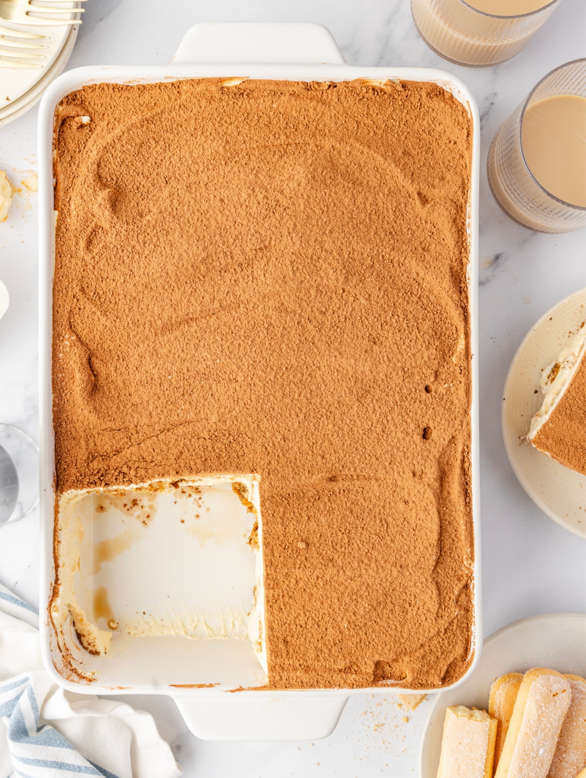 An overhead view of Baileys Irish Cream Tiramisu in a baking dish with a cocoa-dusted surface and one slice lifted out. Baileys Irish Cream Tiramisu in a 9×13-inch dish with a cocoa-dusted top and a single portion removed to show the creamy mascarpone layer underneath.