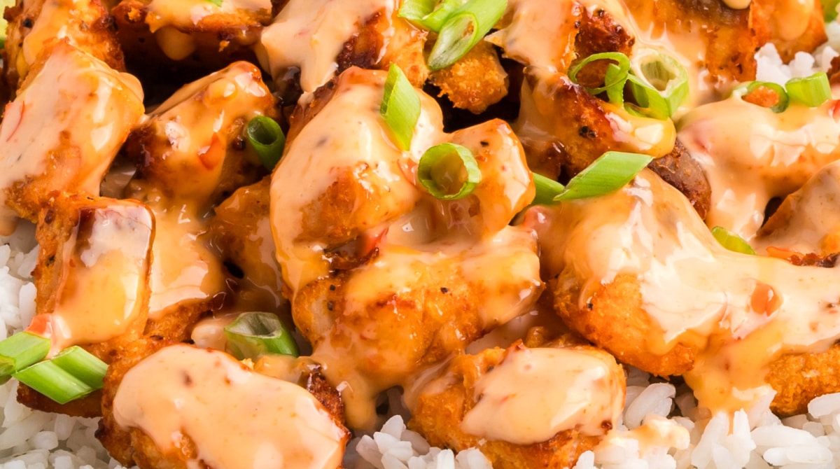 Close-up of crispy salmon bites coated in creamy bang bang sauce and topped with sliced green onions over white rice.