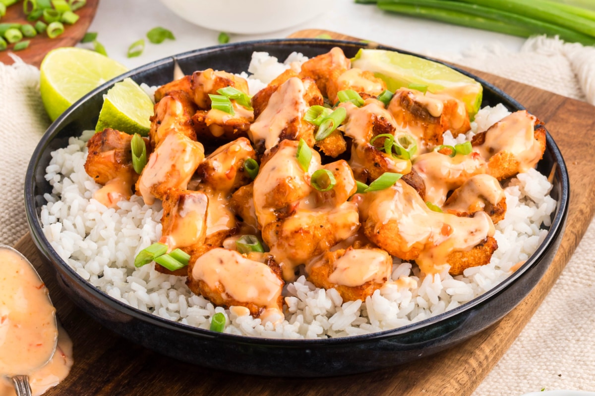 Crispy salmon bites drizzled with creamy bang bang sauce and served over white rice with sliced green onions and lime.