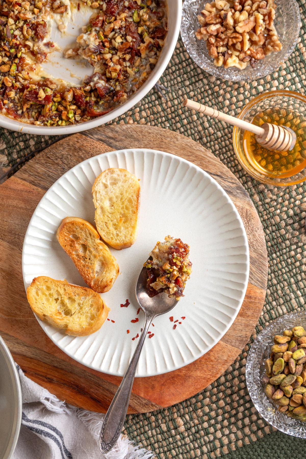 A plated serving of Boursin Bacon Pistachio & Fig Dip with toasted baguette slices and a spoonful showing the creamy, nut-studded texture. A spoonful of Boursin Bacon Pistachio & Fig Dip served on a plate with toasted bread, honey, pistachios, and walnuts.