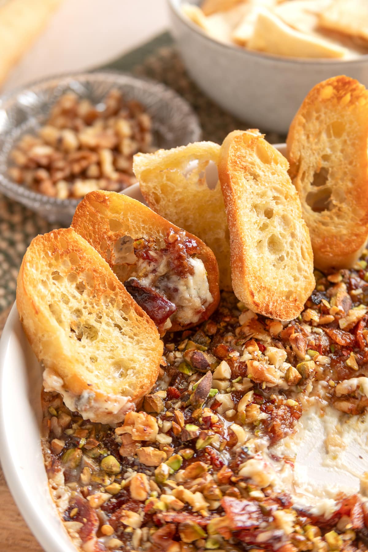 Toasted baguette slices rest in a warm baked Boursin dip topped with crisp bacon, chopped pistachios, walnuts, and fig preserves. Toasted baguette slices standing in a warm baked Boursin dip topped with bacon, fig preserves, pistachios, and walnuts.