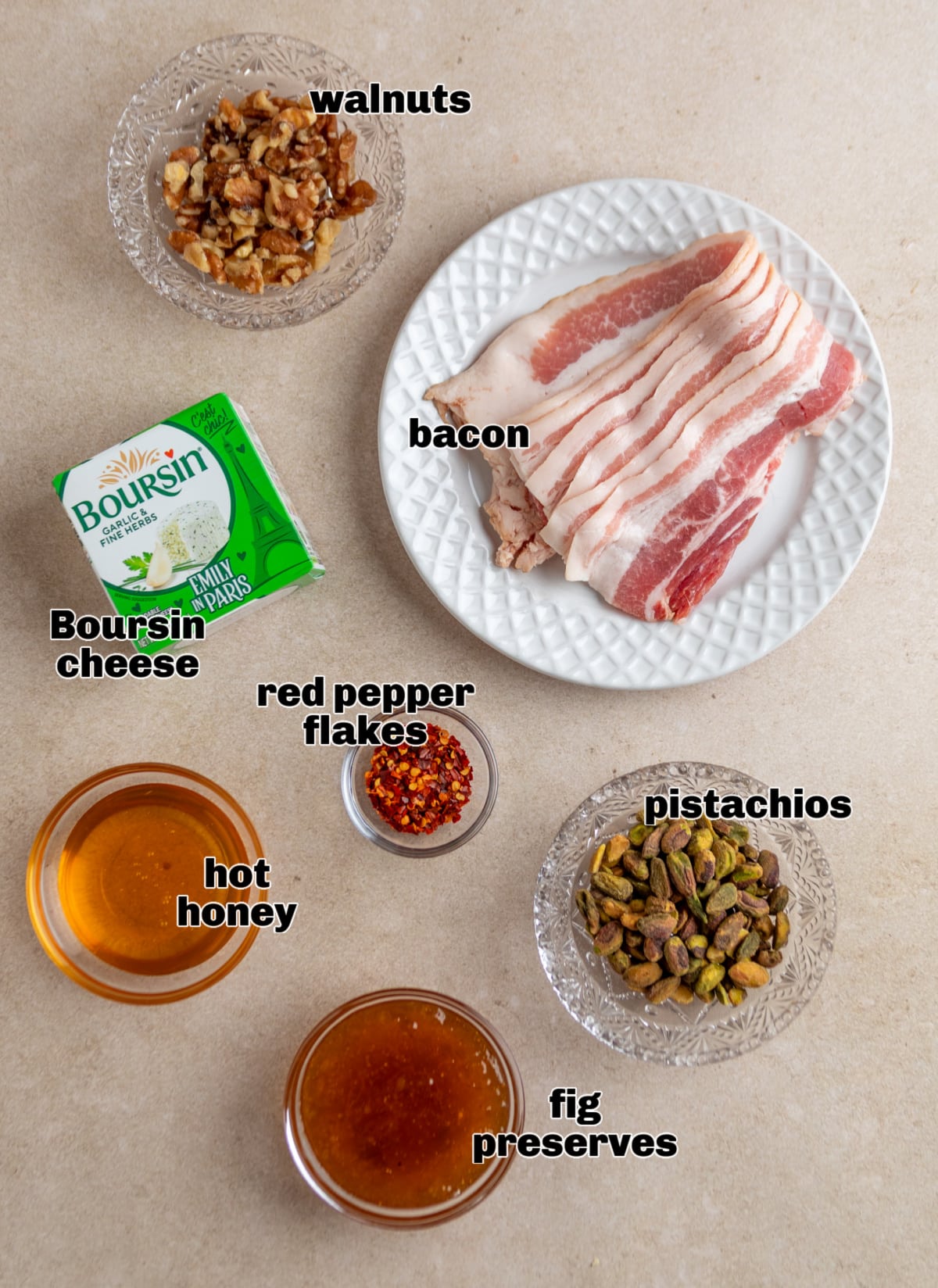 The ingredients for this baked Boursin dip are arranged on the counter, including bacon, Boursin cheese, fig preserves, pistachios, walnuts, red pepper flakes, and hot honey. Ingredients for a baked Boursin dip including Boursin garlic herb cheese, thick-cut bacon, fig preserves, pistachios, walnuts, red pepper flakes, and hot honey.