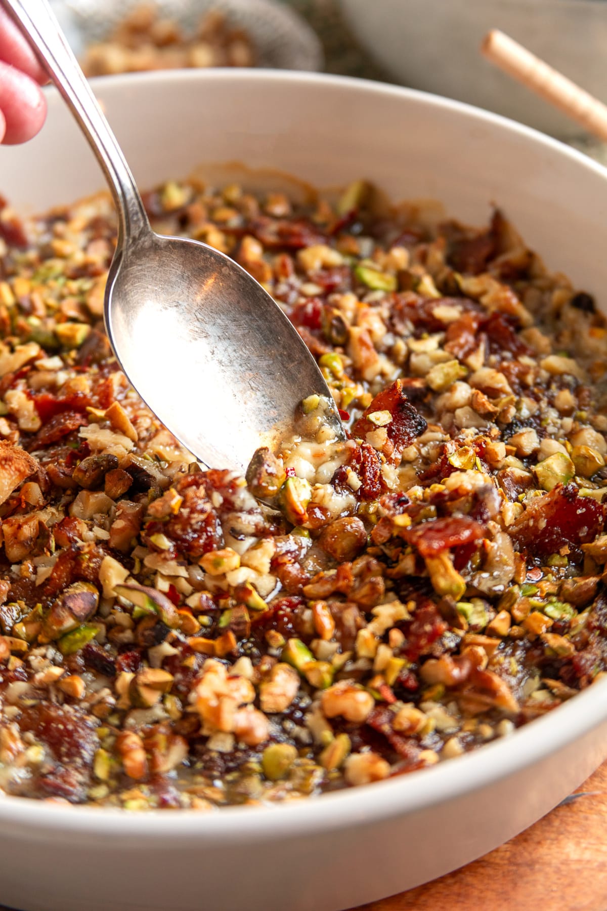 A spoon lifts a scoop of warm baked Boursin dip with crisp bacon, chopped pistachios, walnuts, and a glossy, melted center. A spoon scooping into a baked Boursin dip with bacon, fig preserves, pistachios, and walnuts while the cheese is still soft and melted.