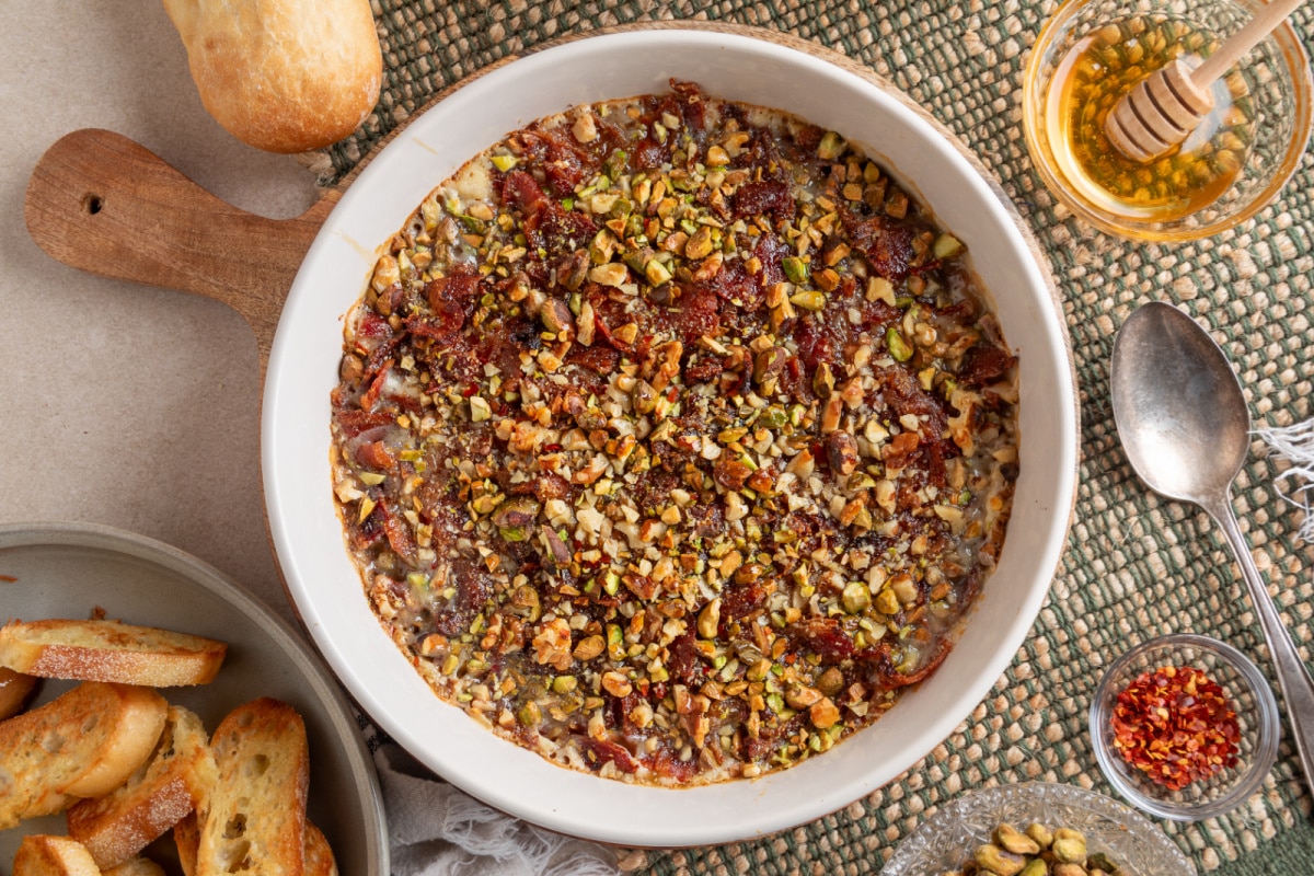 A baked dish of Boursin Bacon Pistachio & Fig Dip fresh from the oven, finished with crisp bacon, chopped nuts, and a drizzle of hot honey. Overhead view of Boursin Bacon Pistachio & Fig Dip baked until bubbly and topped with bacon, pistachios, walnuts, and hot honey.