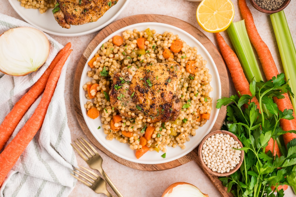 Plated chicken thighs served over pearl couscous with carrots, celery, and herbs, shown with fresh vegetables on the side.