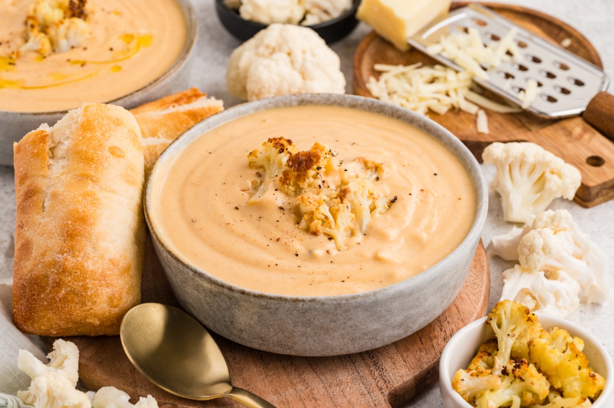 A bowl of Roasted Cauliflower Soup garnished with roasted cauliflower florets and served with bread on a cutting board. Roasted Cauliflower Soup served in a bowl with crusty bread, garnished with caramelized cauliflower florets.