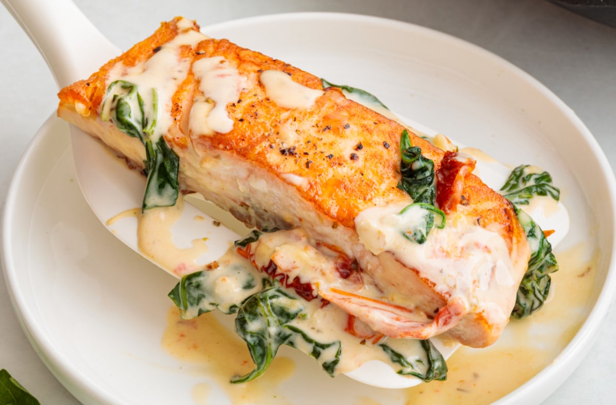 A plated portion of creamy Tuscan salmon served with garlic cream sauce, sun-dried tomatoes, and spinach. Creamy Tuscan salmon lifted from the pan with a spatula, coated in garlic cream sauce with sun-dried tomatoes and spinach.