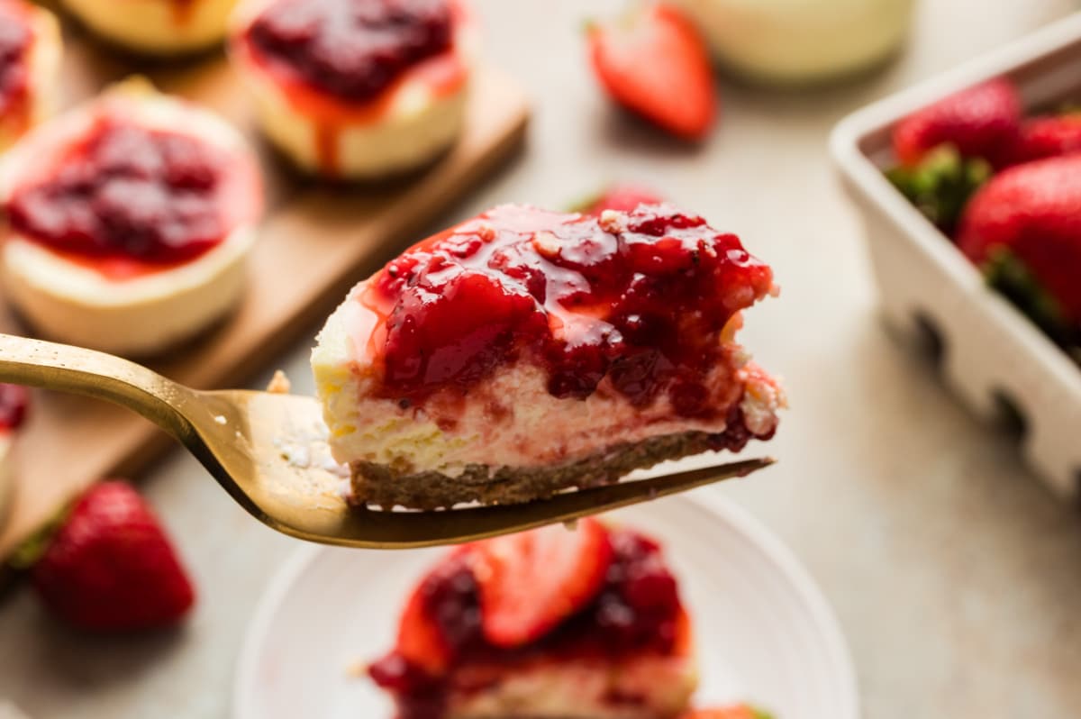 A forkful of mini cheesecake with strawberry topping taken from a fully chilled cheesecake. A fork holding a bite of an individual mini cheesecake with graham cracker crust and fresh strawberry topping, showing the creamy interior and layered texture.