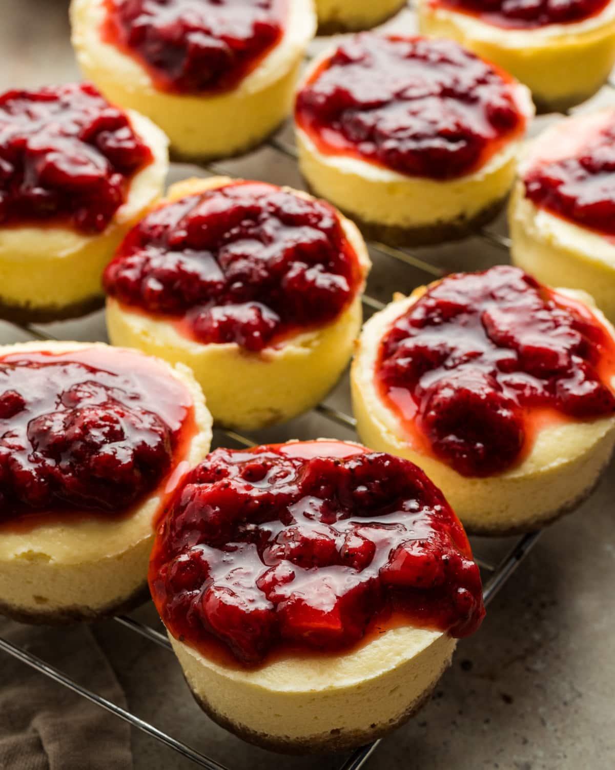 Individual baked mini cheesecakes resting on a cooling rack with strawberry topping set on top. Mini cheesecakes topped with a thick layer of cooked strawberry topping, cooling on a wire rack after baking.