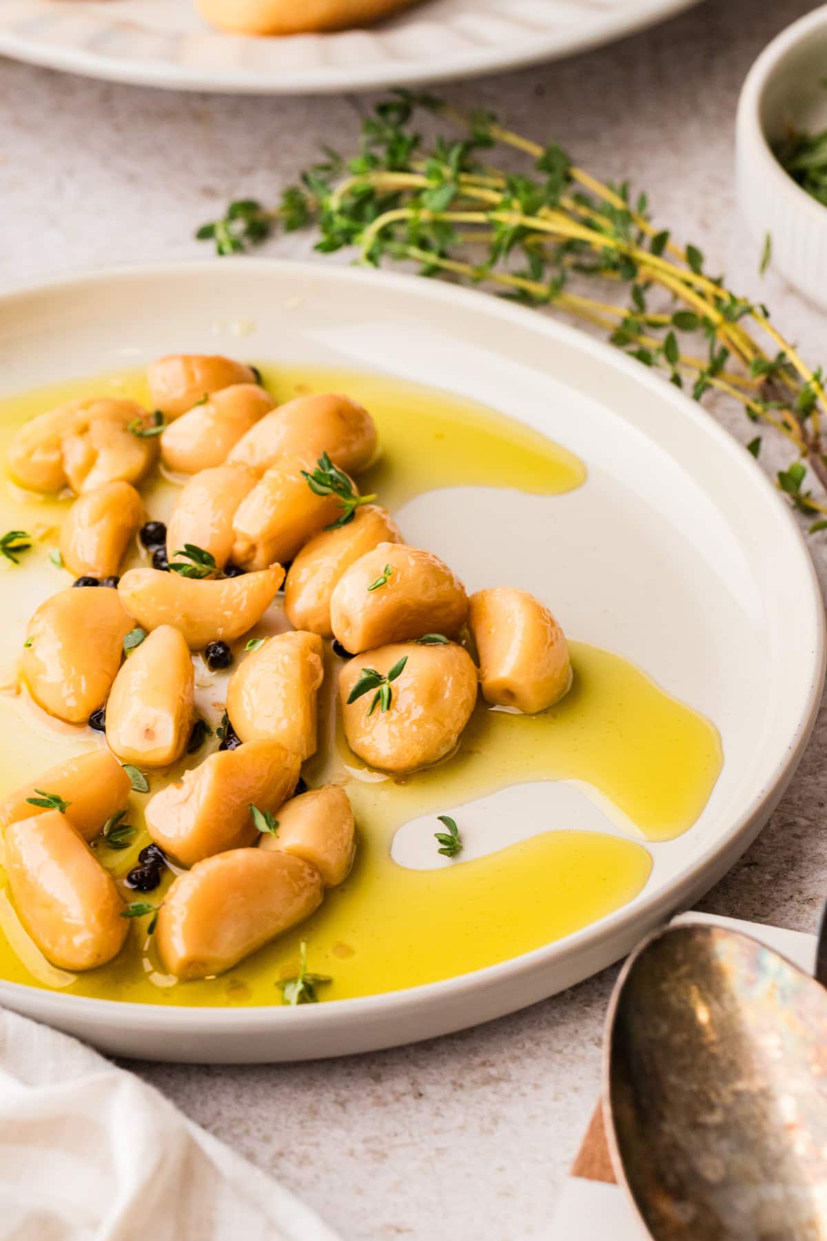 Garlic confit cloves resting in olive oil with thyme and peppercorns on a white plate.