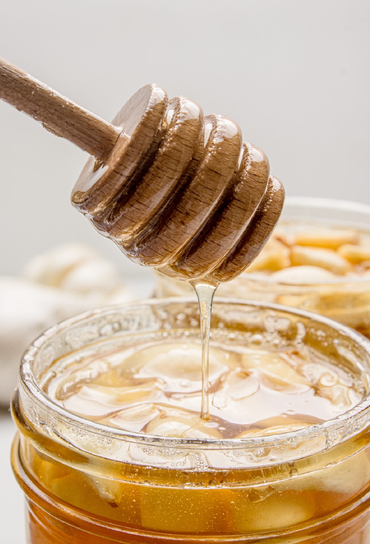 A honey dipper lifting loosened honey from a jar of fermented garlic cloves during the fermentation process. Honey dripping from a wooden honey dipper into a jar of fermented garlic cloves as the honey loosens during fermentation.